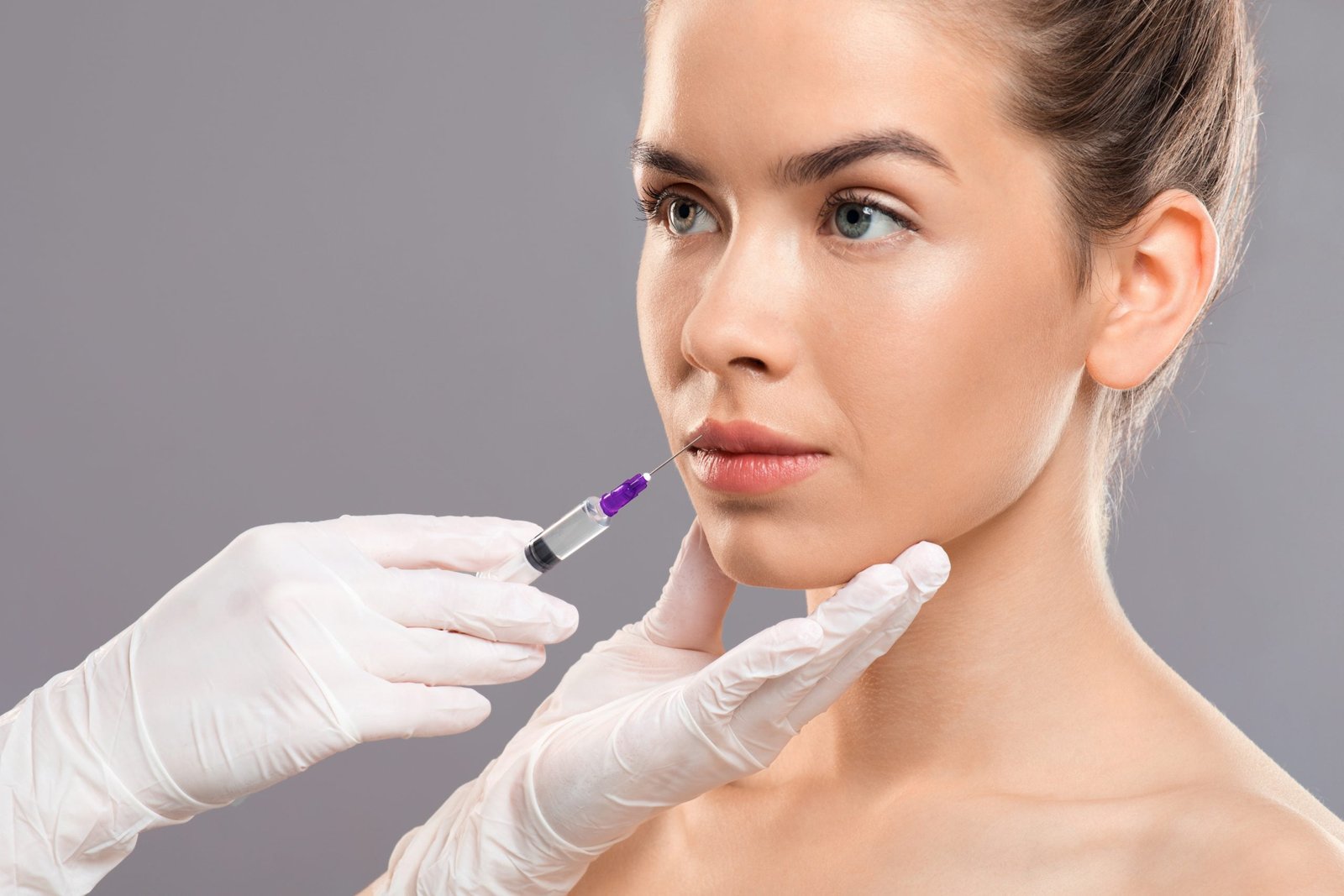 Aesthetic modern medicine concept. Lip injection for young woman over grey studio background, closeup. Unrecognizable cosmetitian injecting lip filler for beautiful lady looking at copy space